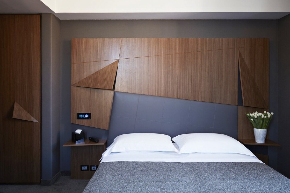 A modern bedroom with a large bed, a nightstand, a control panel, and a vase with flowers.