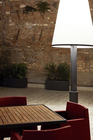 An outdoor seating area with a wooden table and red chairs, a metal lamp post, and potted plants against a brick wall. - The Building (Rome)