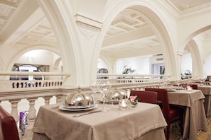 Une salle à manger élégante, avec des tables dressées pour le service, un décor de plafond raffiné et un balcon agrémenté d’une balustrade.
