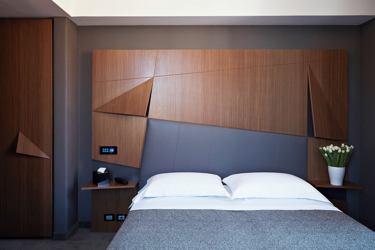 A modern bedroom with a large bed, a nightstand, a built-in desk, and a wall-mounted control panel.