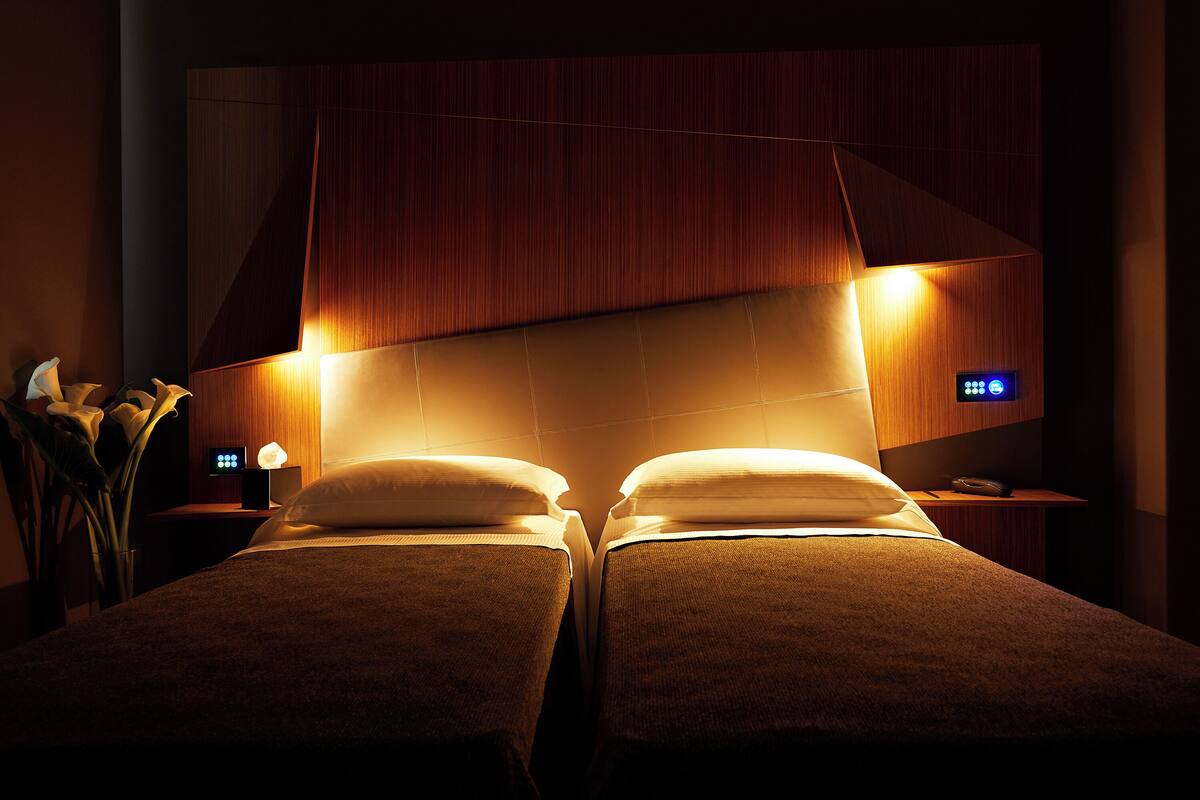 A hotel room with two beds, a nightstand with a lamp, and a wall-mounted control panel.