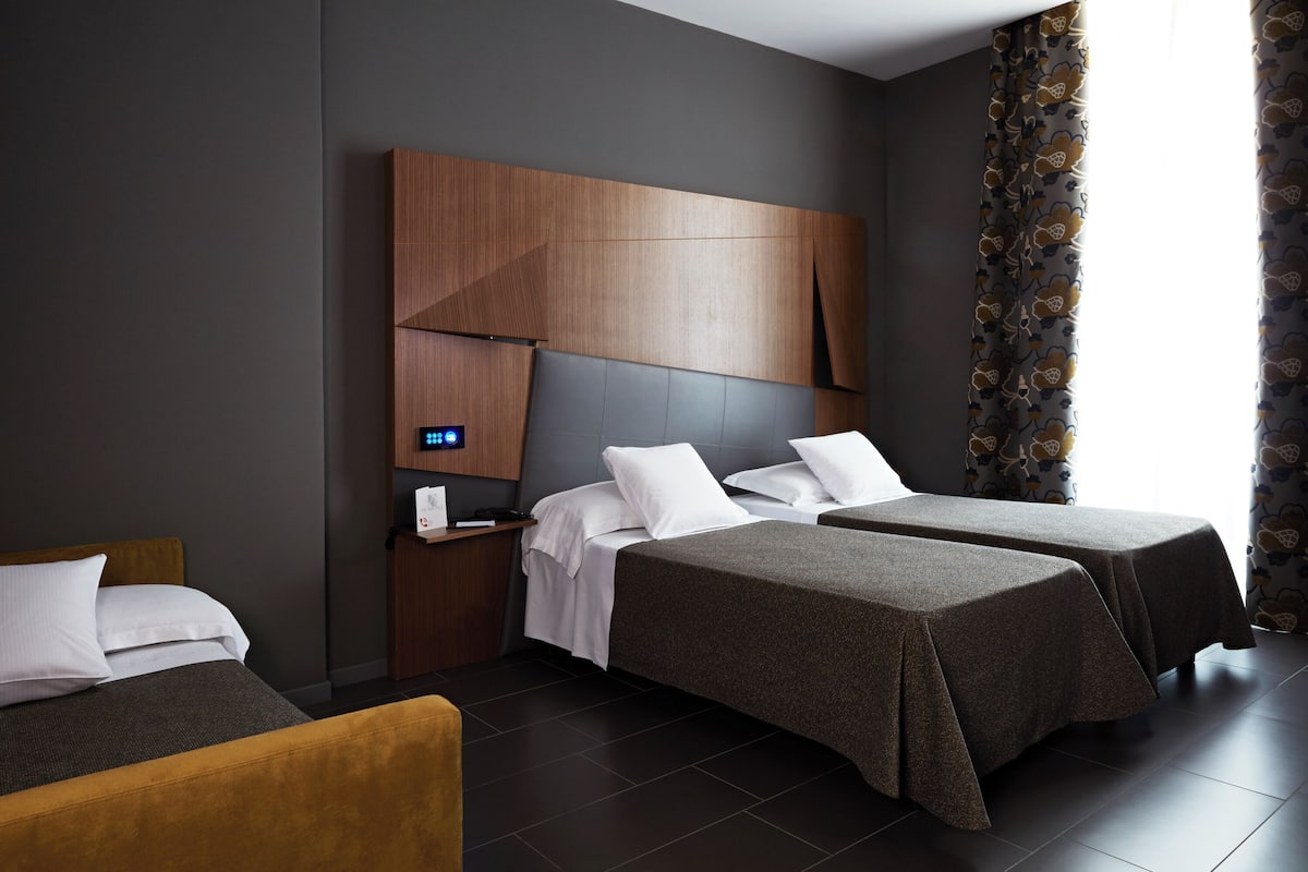 A modern hotel room with two single beds, a wooden headboard, a bedside table with a digital clock, and patterned curtains.