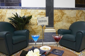A reserved seating area with two black leather chairs, a sign reading "RESERVATO", a martini glass with blue liquid, another with red liquid, a bowl of snacks, and a potted plant. - The Building (Rome)