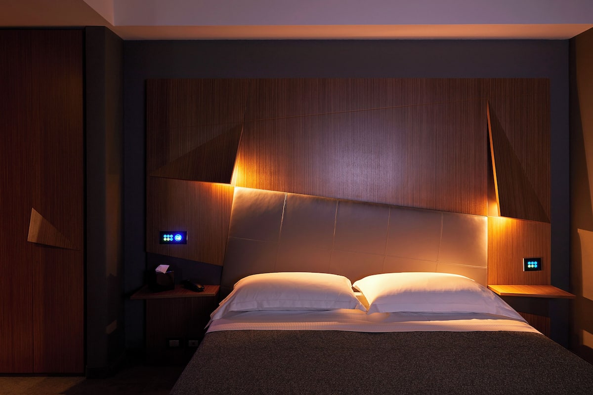 A modern hotel room with a large bed, wooden headboard, bedside tables, and built-in lighting.