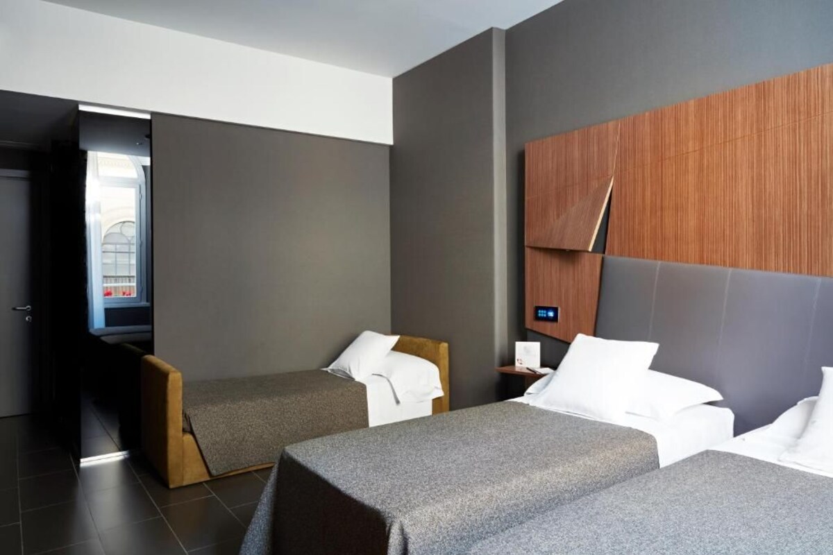 A modern hotel room with two single beds, a wooden headboard, a small bedside table, and a view of a building through an open door.