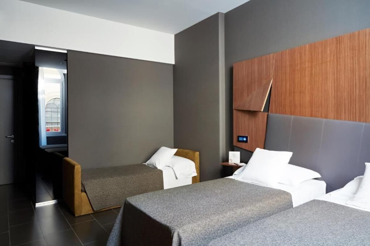 A modern hotel room with two single beds, a wooden headboard, a small bedside table, and a view of a building through an open door.