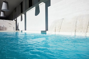 Une piscine agrémentée d’une cascade, entourée d’une architecture moderne avec de grandes fenêtres et des luminaires suspendus.