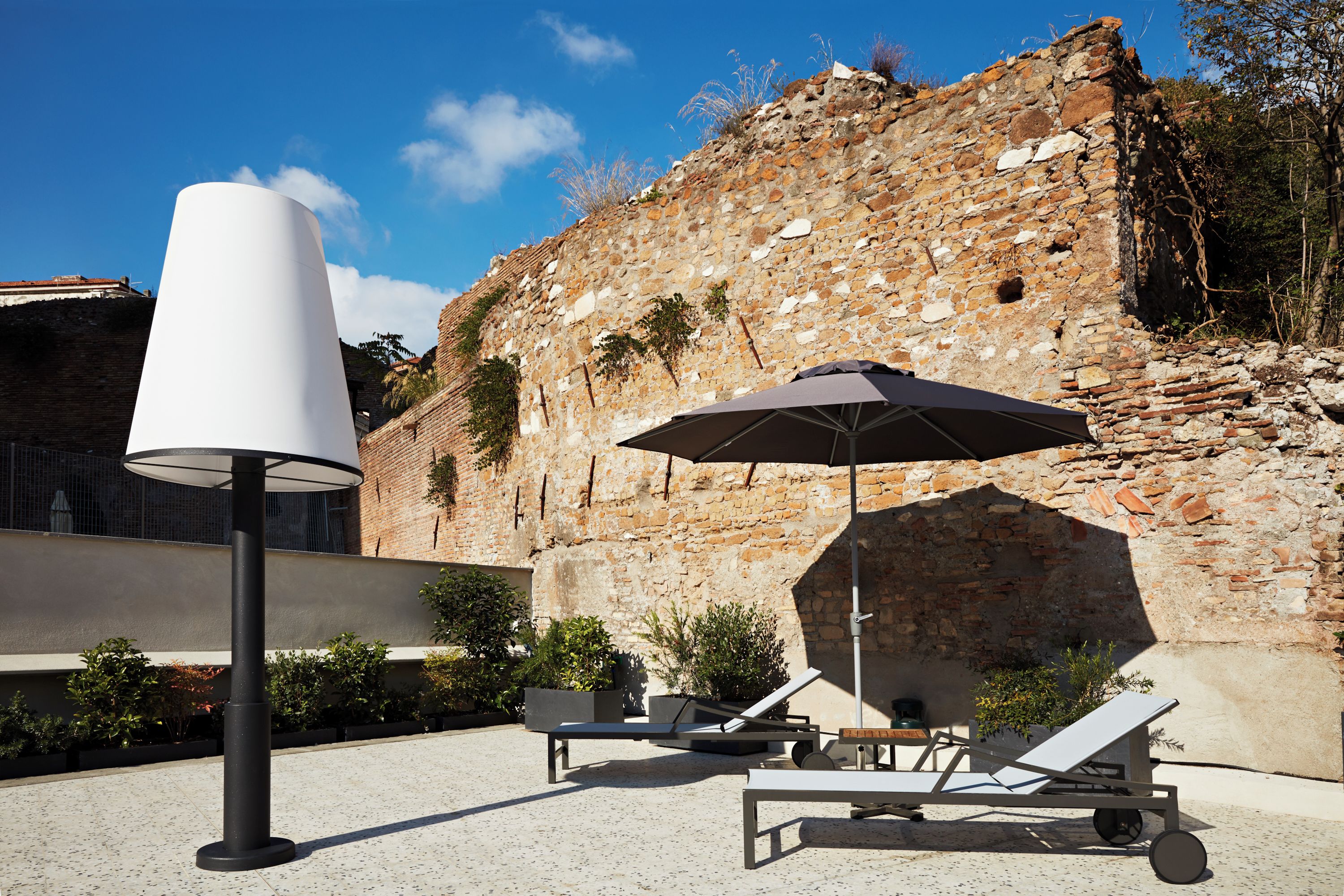 Un patio con muro de piedra, sillones de descanso, un paraguas grande y un farol moderno.