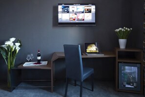 Une chambre d’hôtel équipée d’un bureau, d’un ordinateur portable, d’un téléviseur fixé au mur, d’une chaise, d’un vase avec des fleurs et d’un petit réfrigérateur.