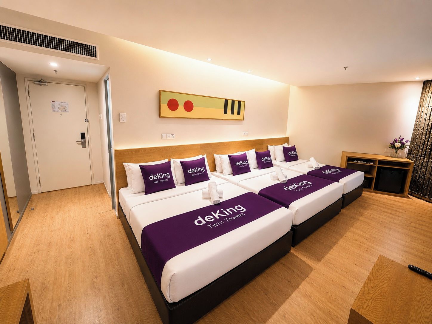A hotel room with two beds, each with a purple bedspread and pillows labeled 'deKing Twin Towers.'
