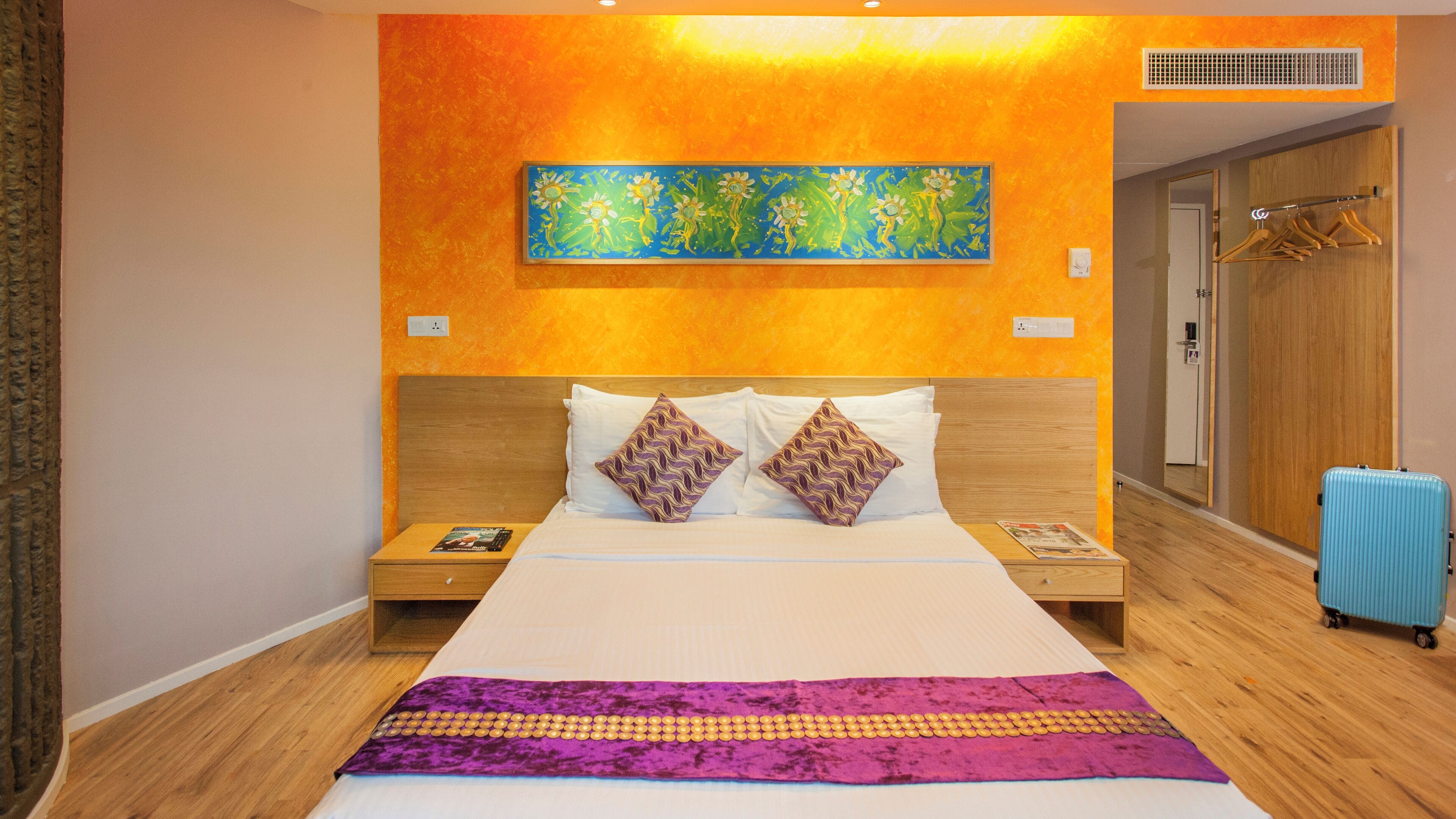 Hotelkamer met een groot bed, een houten hoofdeinde, twee nachtkastjes, een blauwe koffer en een kleurrijk schilderij aan de muur.