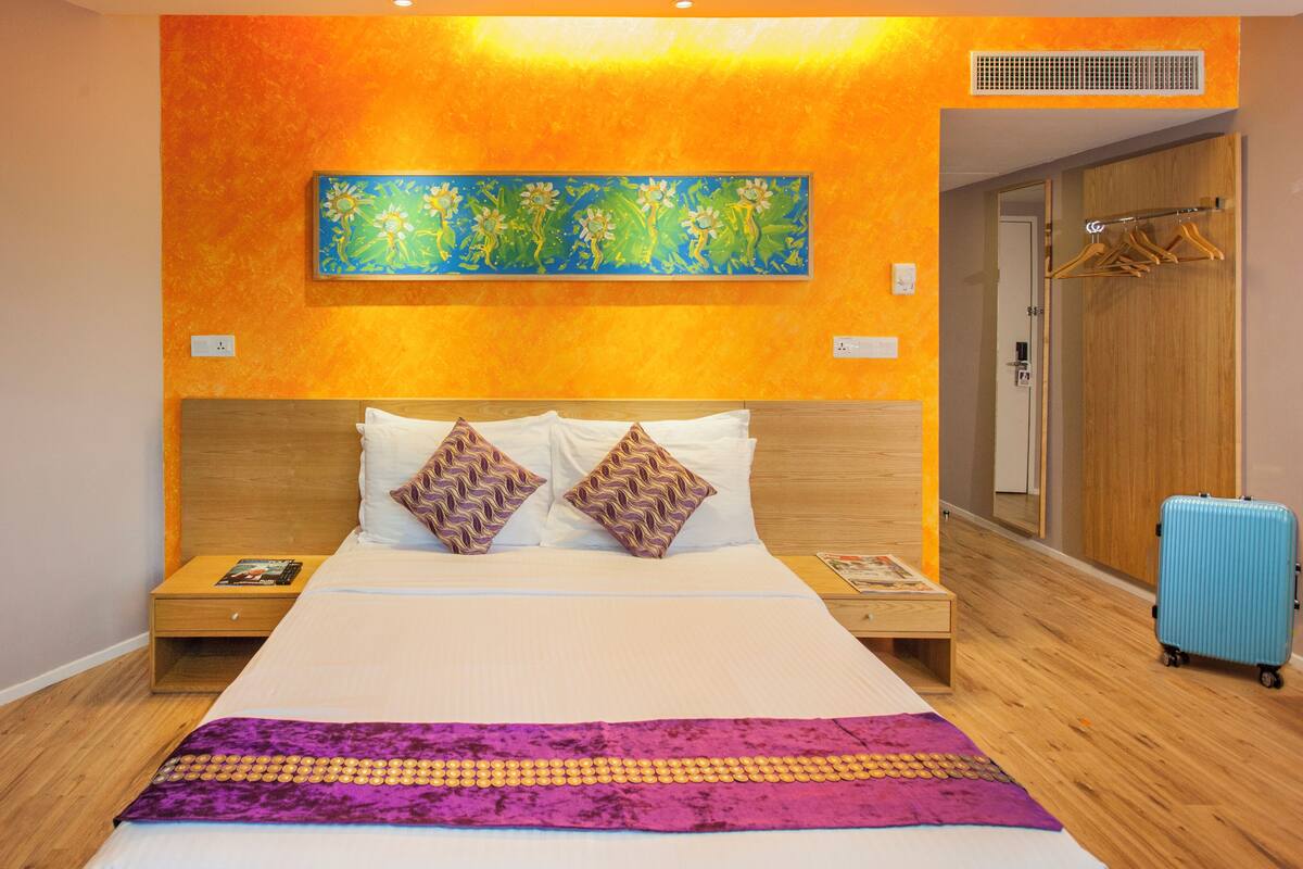 Ein Hotelzimmer mit einem großen Bett, einem Holzkopfteil, zwei Nachttischen, einem blauen Koffer und einem farbenfrohen Bild an der Wand.