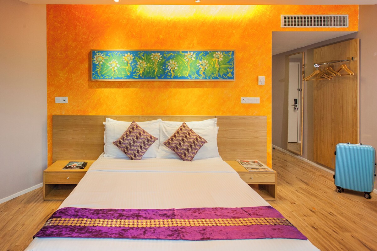 A hotel room with a large bed, wooden headboard, two nightstands, a blue suitcase, and a colorful painting on the wall.