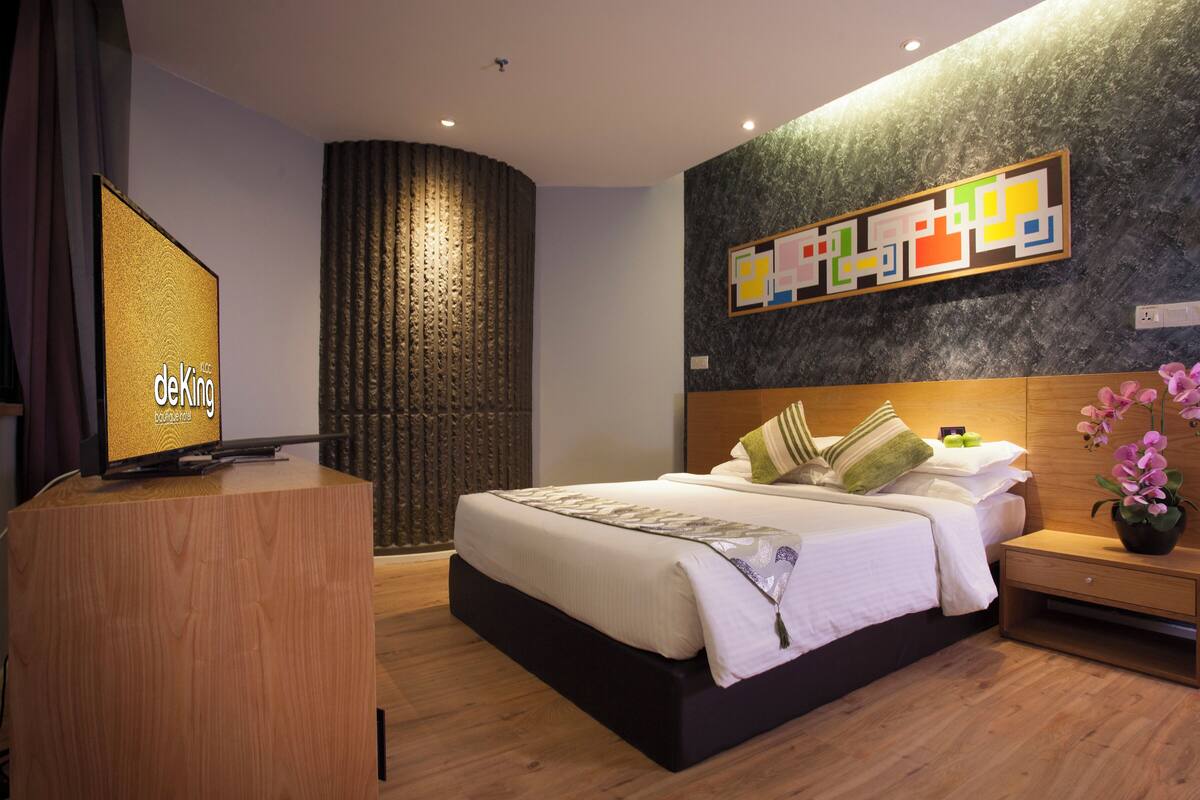 Ein modernes Hotelzimmer mit einem großen Bett, einem Holztisch, einer Wand mit Dekoration und einem Fernseher, der 'deKing' anzeigt.