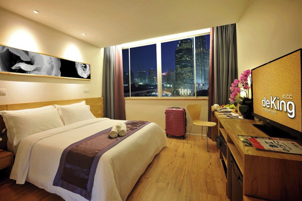 A hotel room with a bed, a desk, a TV, a window with a city view, and a painting on the wall.