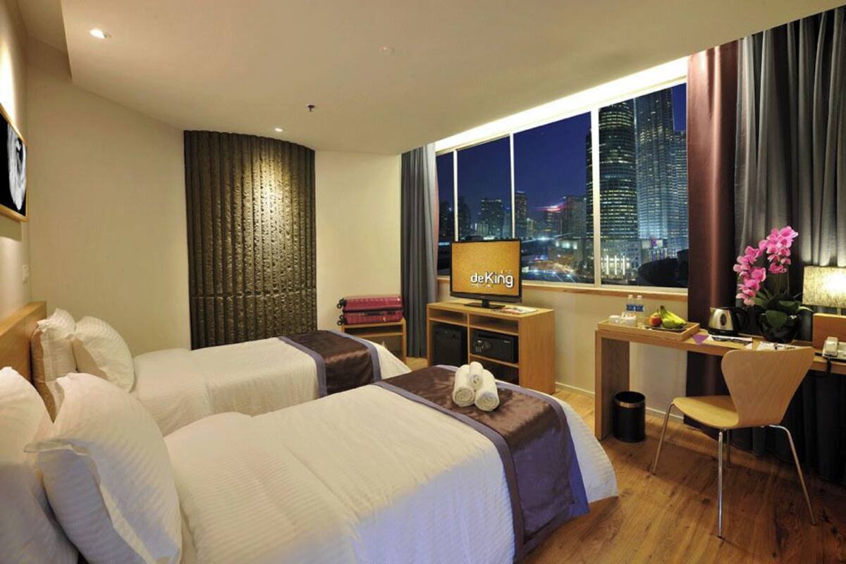Ein Hotelzimmer mit zwei Betten, einem Schreibtisch, einem Sessel, einem Fernseher und Blick auf eine beleuchtete Stadt bei Nacht.