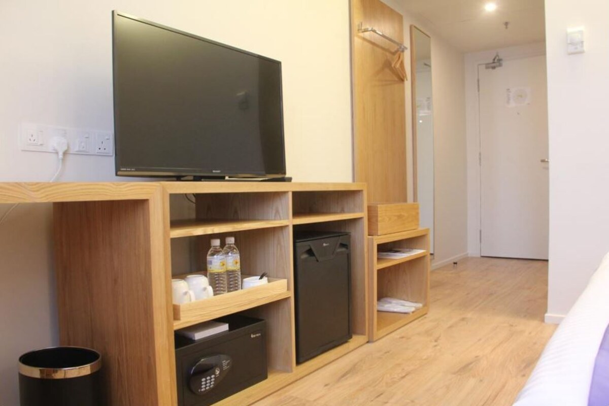A modern hotel room with a flat-screen TV on a wooden shelf, a compact refrigerator, and a trash bin.