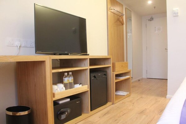 Una habitación de hotel moderna con una televisión de pantalla plana sobre un estante de madera, un refrigerador pequeño y un cesto de basura.