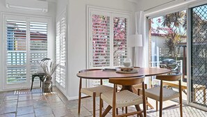 A dining area with a round table and four chairs, a vase with dried flowers, and a window with white shutters. - Dora Cottage • Fireplace + Firepit • Walk to CBD (Orange)