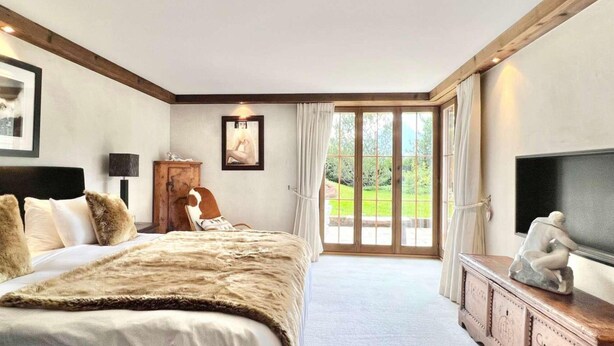 A bedroom with a large bed, a wooden headboard, a television, and a view of a garden through a glass door. - 7-bedroom Chalet With Private Pool (Saanen)