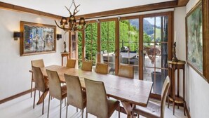 A dining area with a large table and chairs, large windows with a view of greenery, and a chandelier. - 7-bedroom Chalet With Private Pool (Saanen)