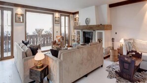 A spacious living room with a large sofa, a fireplace, and a view through large windows. - 7-bedroom Chalet With Private Pool (Saanen)