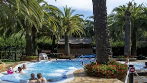 Zona de hidromasaje con gente relajándose, rodeada de palmeras y una piscina con suelo de piedra.