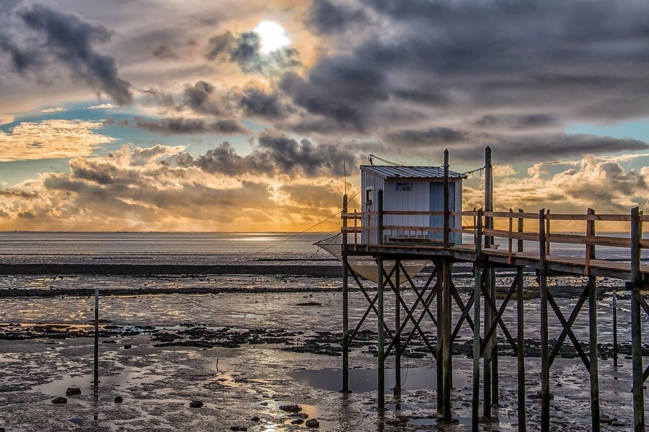 A pier with a small building at the end, overlooking a beach with a sunset in the background.