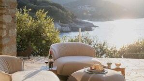 A stone-floored patio with two beige sofas and a small table with a glass of wine and a bottle. A scenic view of a coastal landscape with mountains and greenery is visible through the open space. - Villa Mare di Spartaia (Marciana)