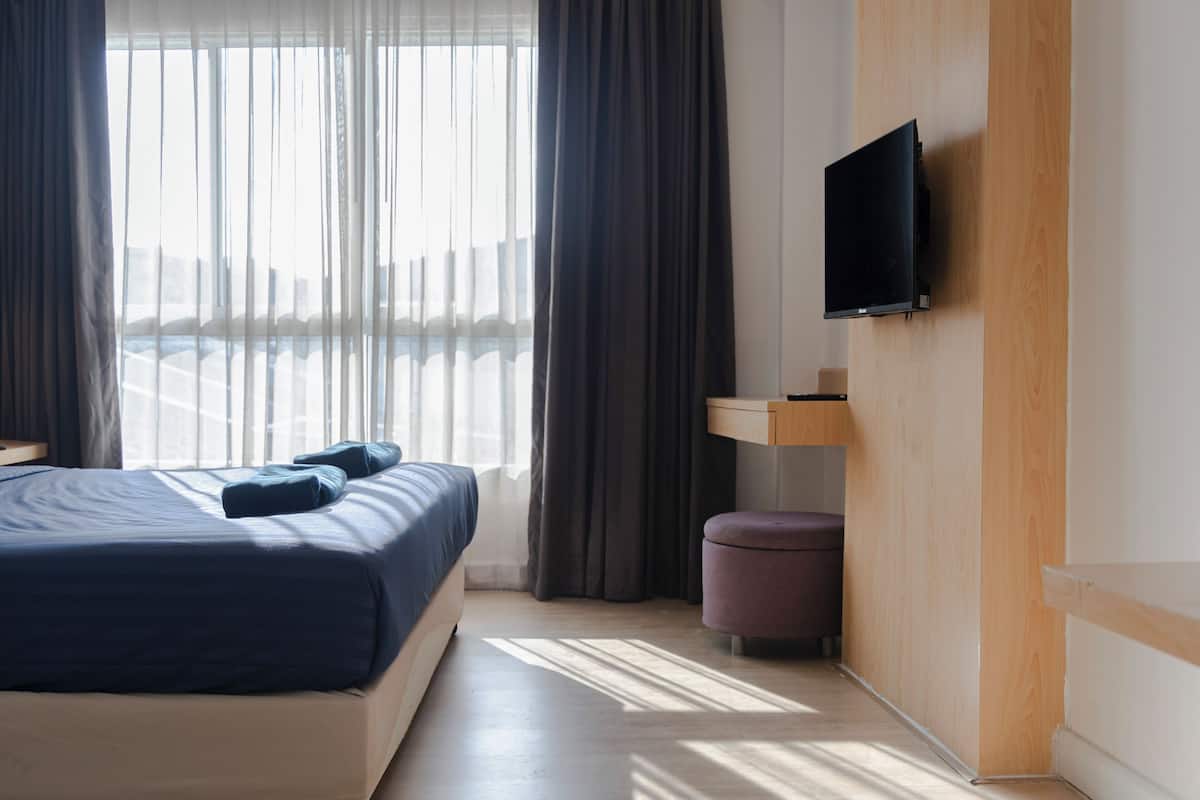 A modern hotel room with a large bed, a flat-screen TV mounted on the wall, a small wooden side table, and a view of the city through sheer curtains.