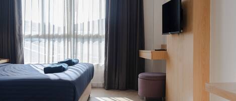 A modern hotel room with a large bed, a flat-screen TV mounted on the wall, a small wooden side table, and a view of the city through sheer curtains.