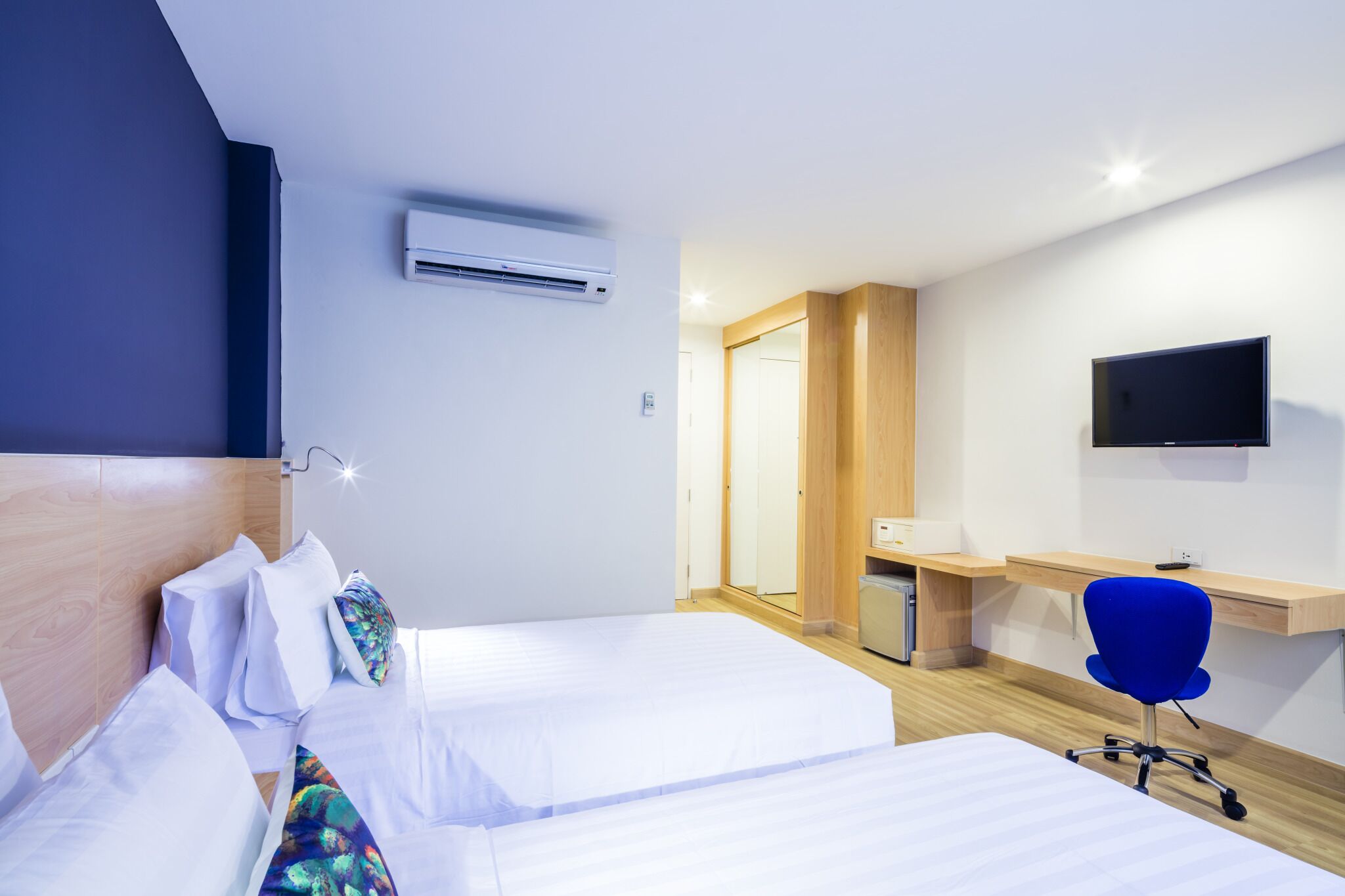 A hotel room with two beds, a desk, a chair, a TV, and an air conditioning unit.
