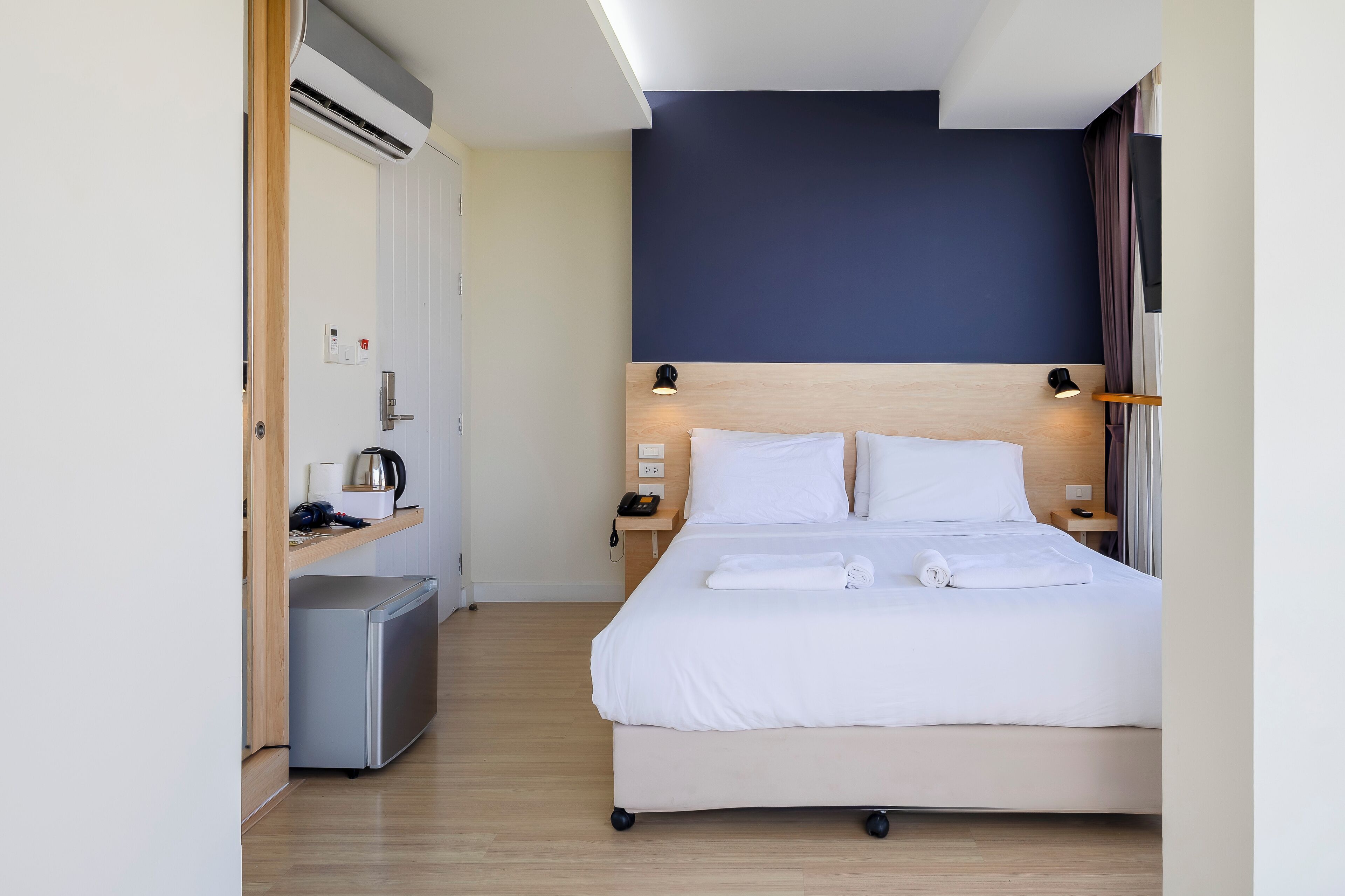 A hotel room with a bed, bedside tables, a small desk, a mini-fridge, and a ceiling-mounted air conditioner.