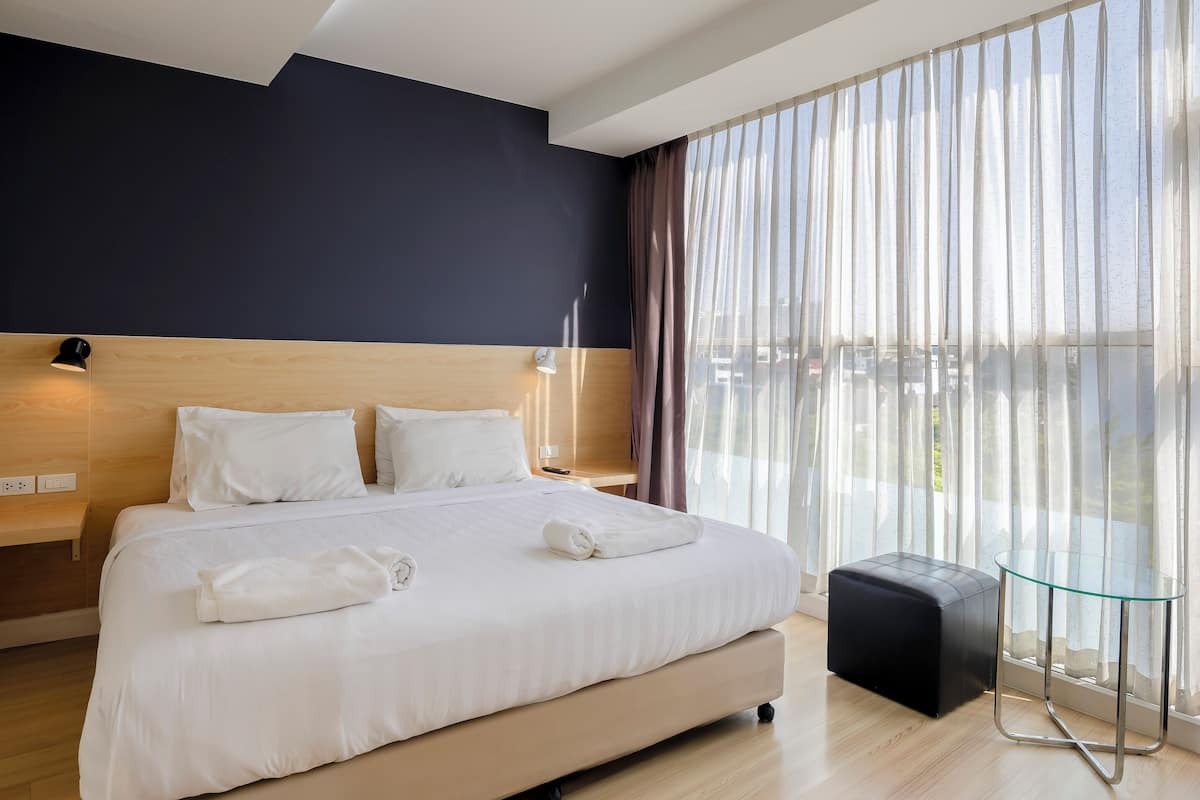 A modern hotel room with a large bed, wooden floor, and a view through sheer curtains.