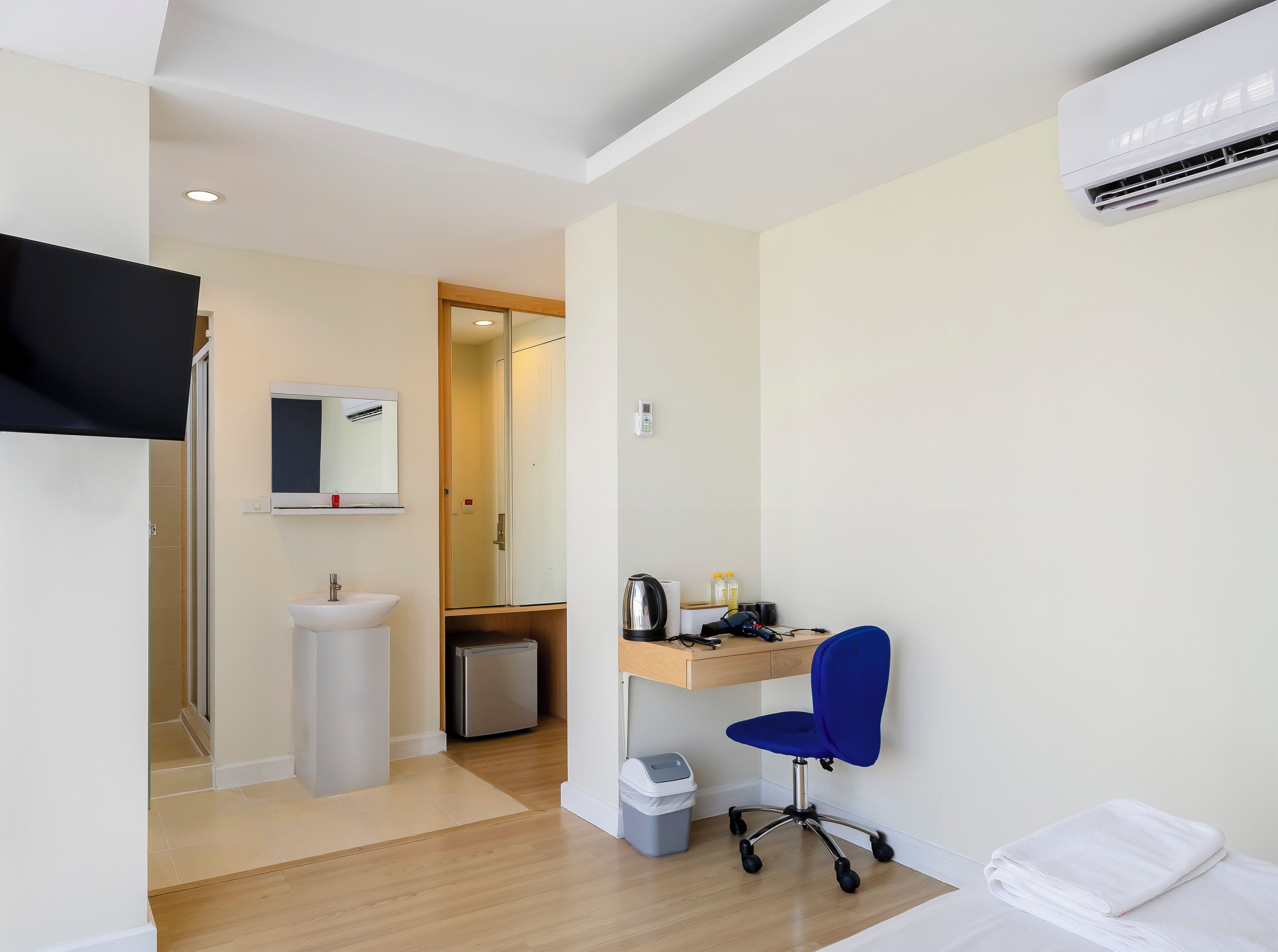 A hotel room with a bed, a desk with a chair, a sink, a mirror, a small refrigerator, and an air conditioning unit.