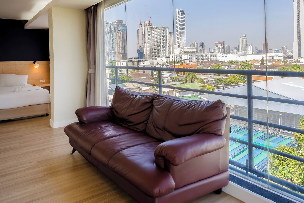 A modern hotel room with a large window, a leather sofa, a bed, and a city view.