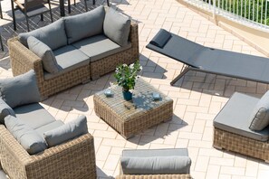Ein Rattansitzgruppe mit Kissen, dazu ein passender Hocker und eine Sonnenliege auf einer gefliesten Terrasse.