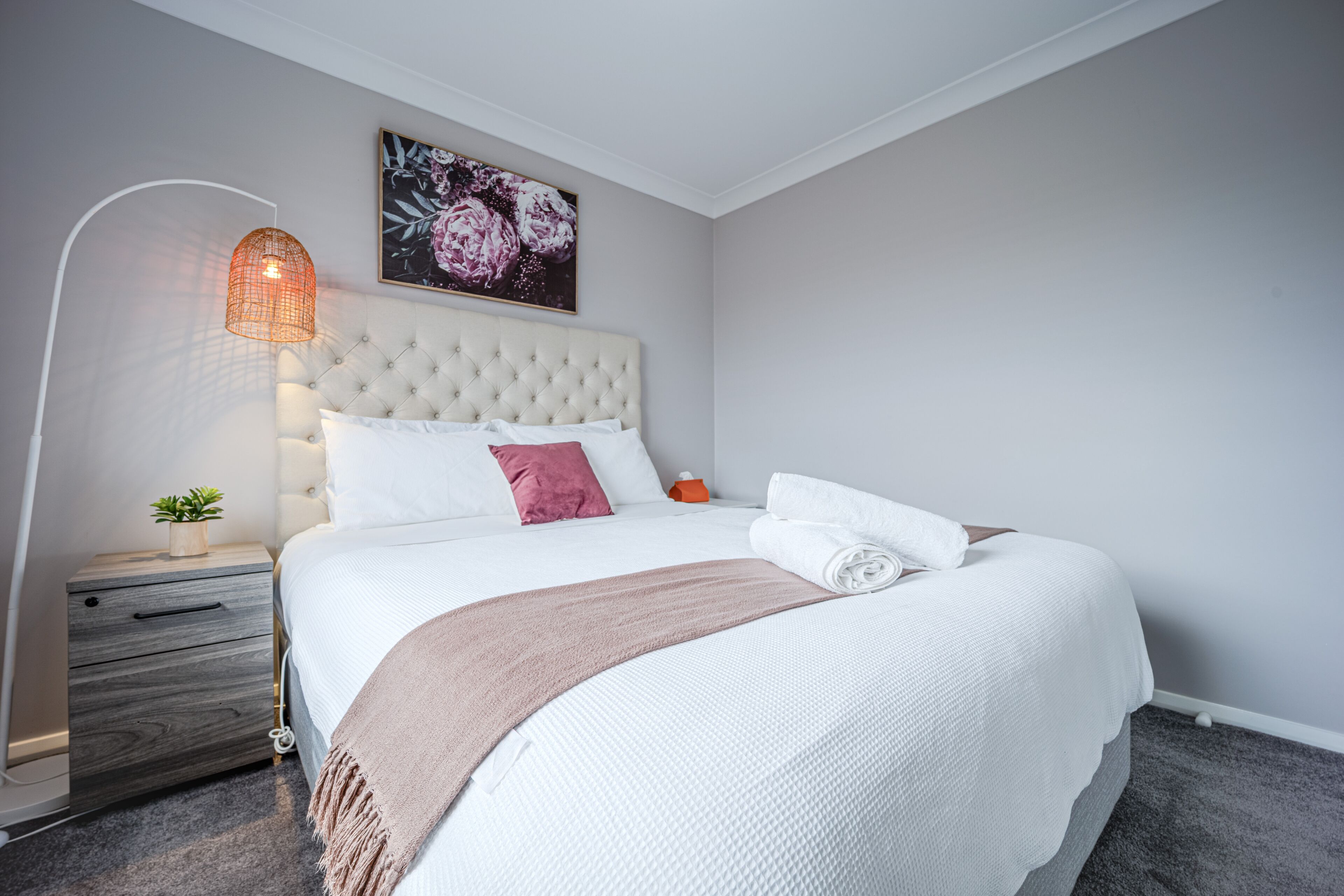A neatly made bed with a white and pink pillow arrangement, a beige throw blanket, and a bedside lamp.