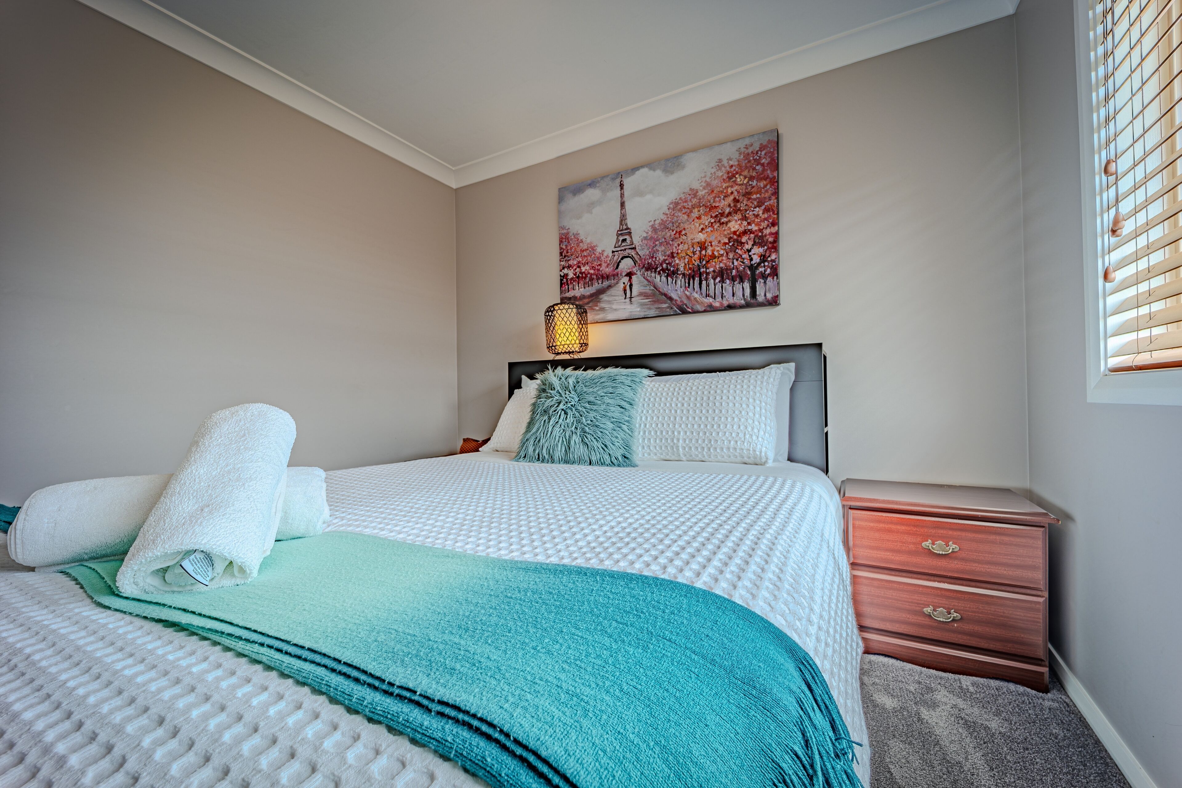 Een net opgemaakt bed met een wit-blauwe deken, een klein nachtkastje met een lamp, en een schilderij van de Eiffeltoren aan de muur.