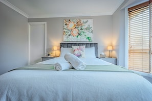 Ein ordentlich bezogenes Bett mit einem zusammengerollten Handtuch, ein Blumenbild über dem Bett, zwei Nachttischlampen und ein Fenster mit Jalousien.