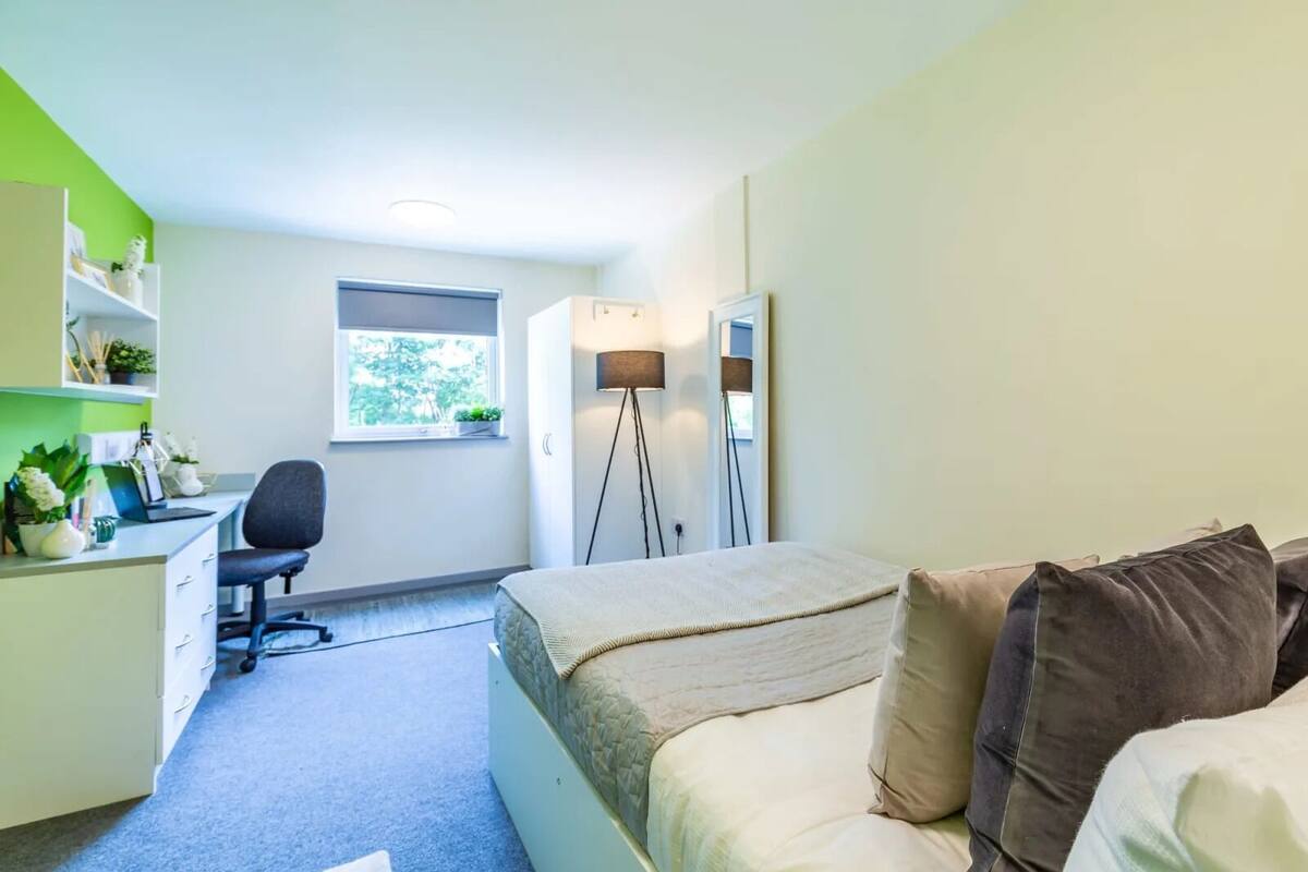 Une chambre à coucher de taille modeste, comprenant un lit, une table de chevet et un bureau avec une chaise. Une fenêtre laisse entrer la lumière naturelle et une lampe sur pied est également présente.