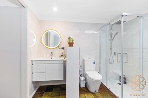 Bathroom features a white vanity, round mirror, toilet, and glass-enclosed shower area.