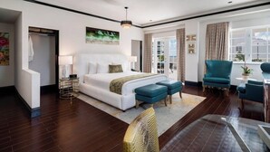 Ein modernes Hotelzimmer mit einem großen Bett, zwei blauen Hocker, einem blaugrünen Sessel, einem kleinen Tisch und Blick ins Freie.