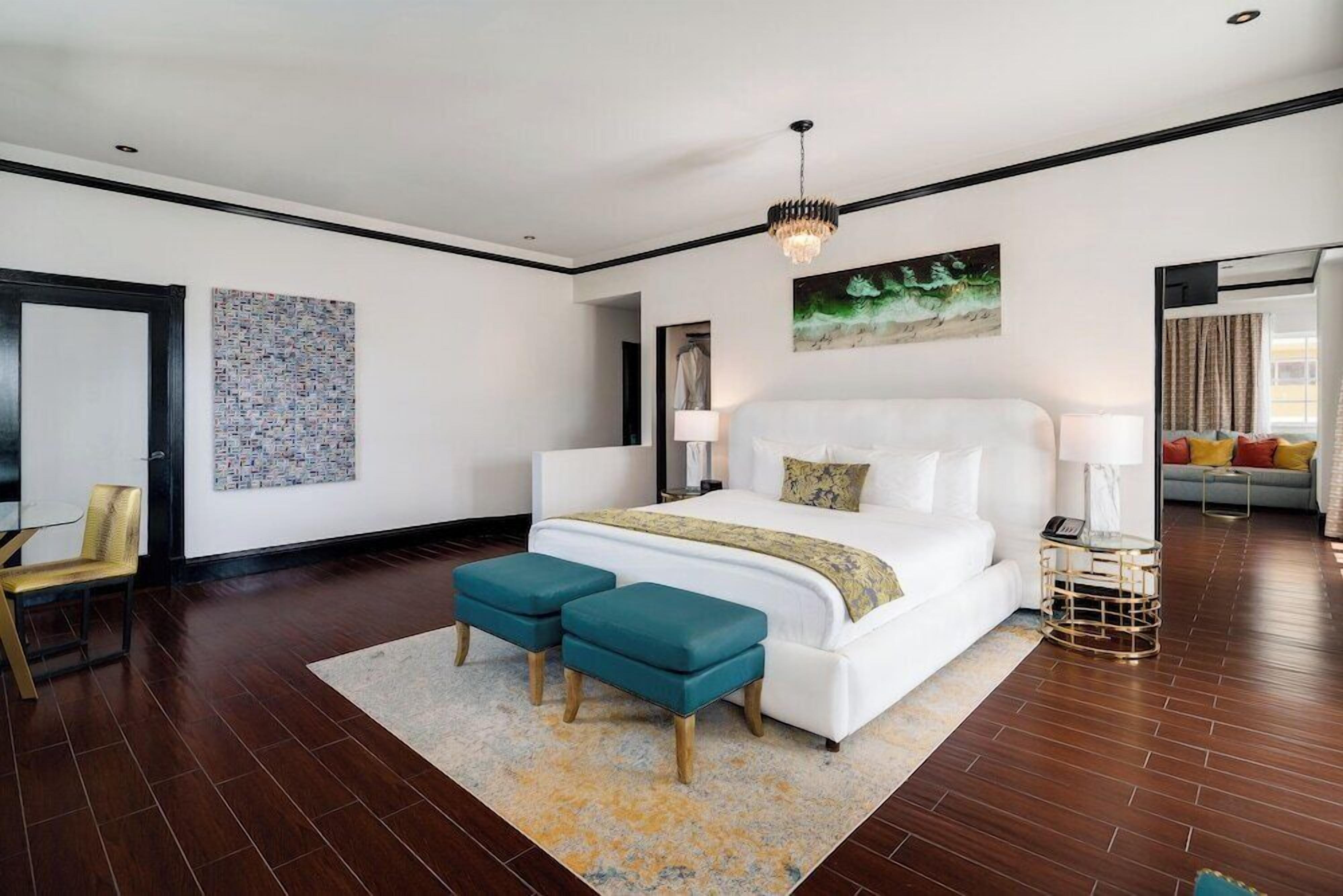 A modern bedroom with a large bed, two matching ottomans, a chandelier, and a painting on the wall.