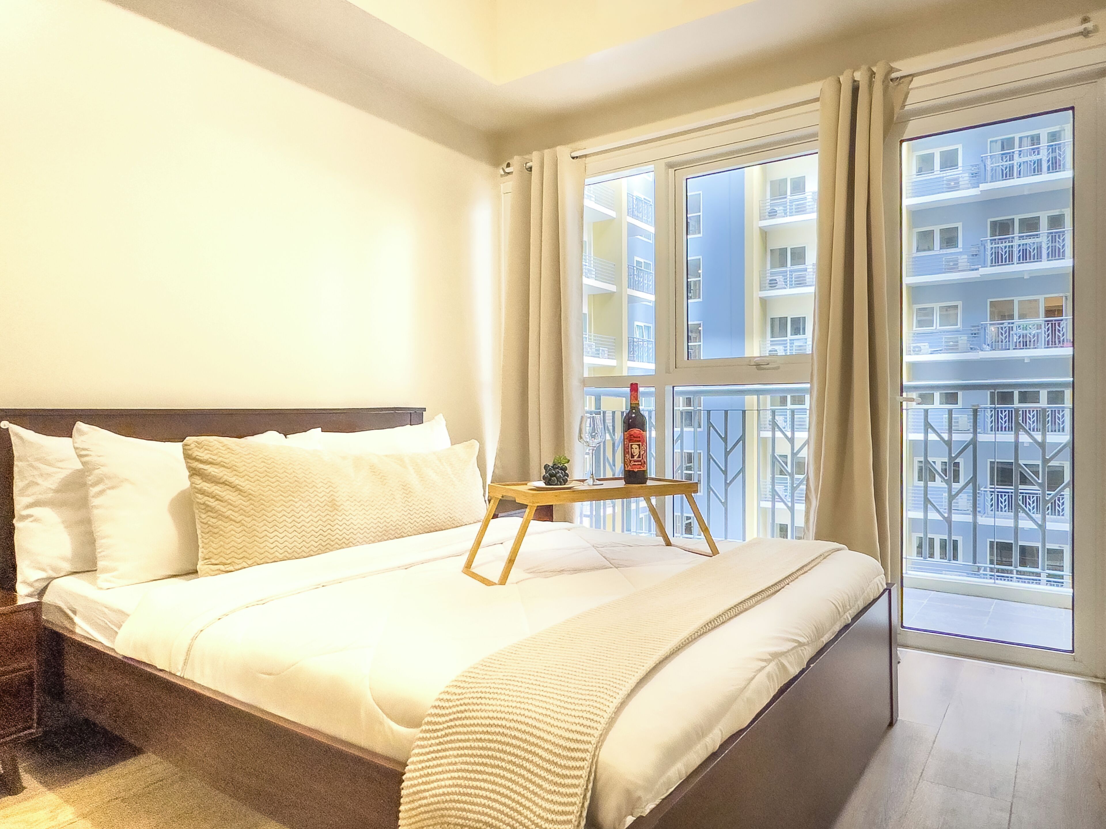Une chambre d’hôtel avec un lit, une petite table sur laquelle se trouvent une bouteille et un verre, et une vue sur un immeuble doté de balcons.