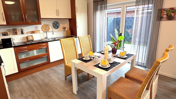 Une cuisine moderne avec des meubles blancs, une table à manger pour quatre personnes et une vue sur l’extérieur grâce à de grandes fenêtres.