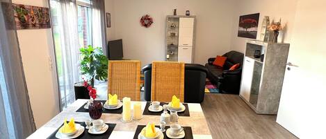 Ruokailutila, jossa on neljälle katettu ruokapöytä, sohva, kaappi ja taustalla televisio.