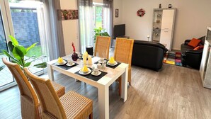 Un coin repas avec une table dressée pour quatre personnes, entouré de chaises en osier. Un téléviseur est posé sur un meuble à droite, et un canapé avec des coussins se trouve au fond de la pièce.