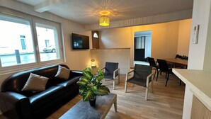Ein modernes Wohnzimmer mit einem schwarzen Ledersofa, einem Couchtisch aus Holz und einer Topfpflanze. Es gibt einen Essbereich mit Tisch und Stühlen sowie eine Küchenzeile mit Herd.