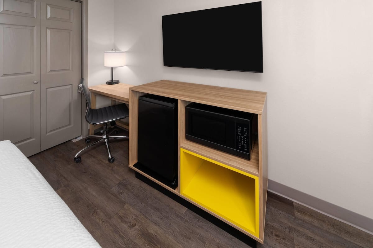 Habitación de hotel con un escritorio de madera, un refrigerador negro, un compartimento de almacenamiento amarillo, una silla negra y un televisor de pantalla plana montado en la pared.